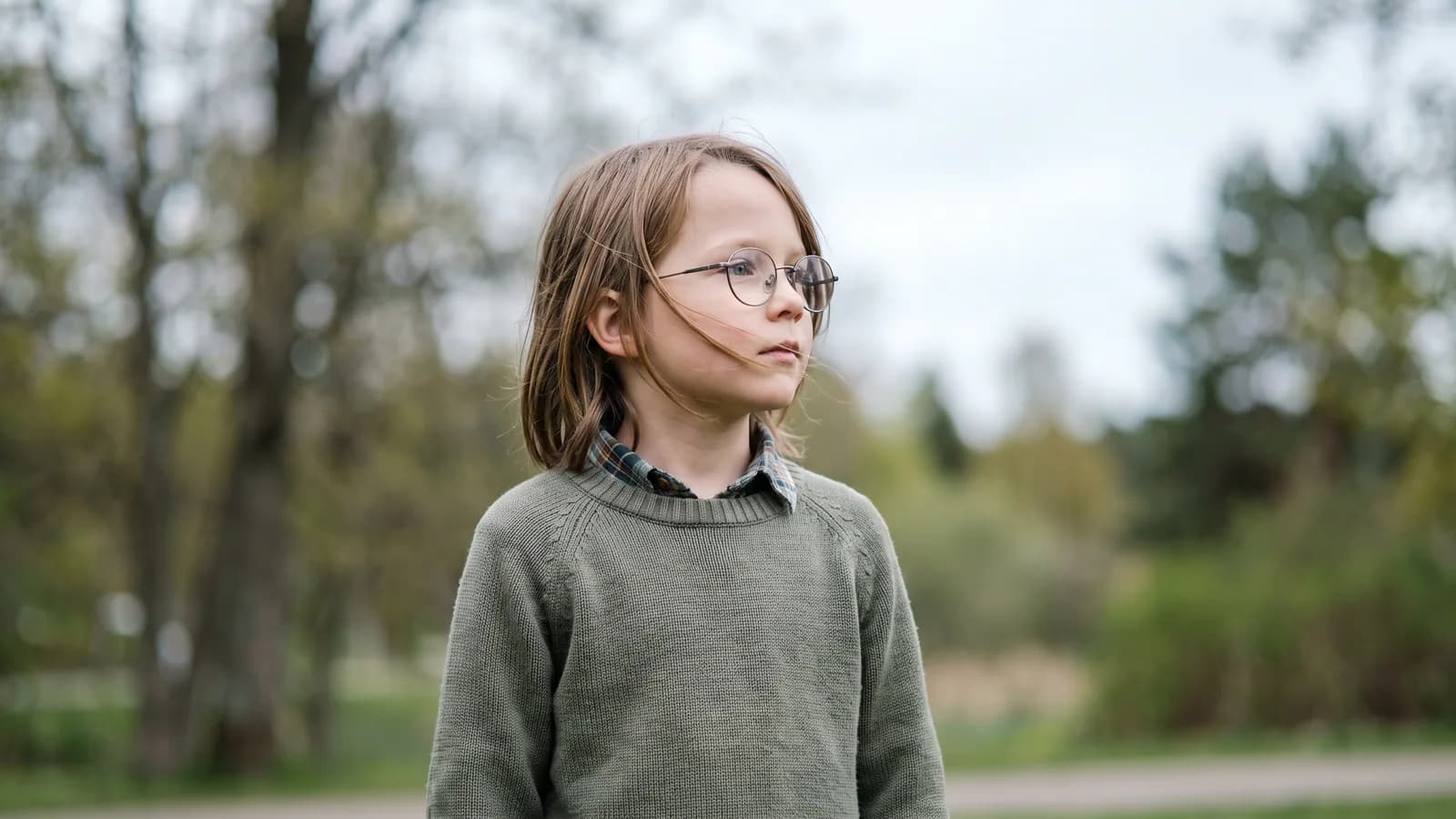 Et barn i skolealder med briller står ute i et lyst nordisk parklandskap og ser nysgjerrig mot horisonten