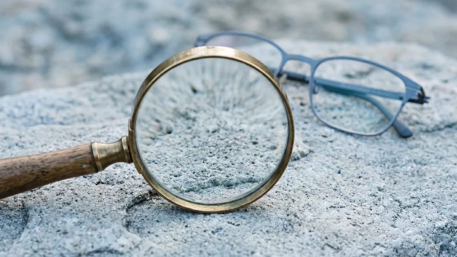 Et forstørrelsesglass som ligger på en kjølig grå steinflate, med et par tynne briller ute av fokus i bakgrunnen