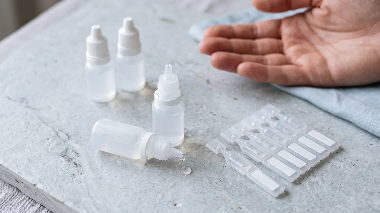 Flere umerkede øyedråpeflasker og en stripe endose-pipetter på en kjølig lys steinflate, med en dråpe på tuppen