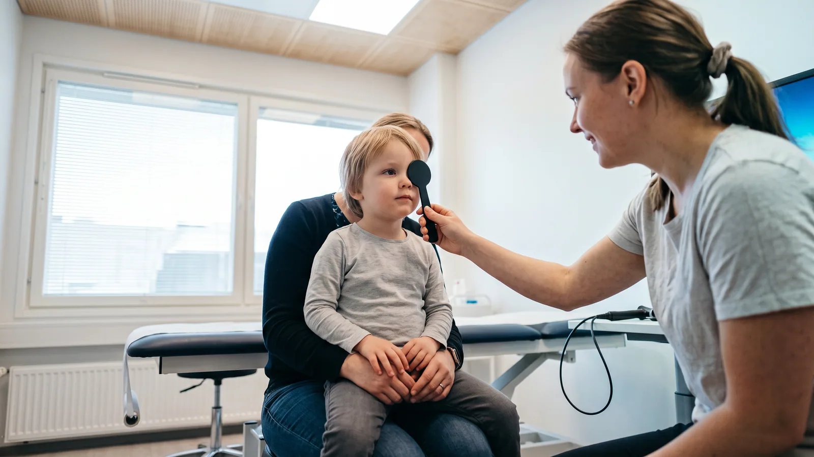 En forelder lyser forsiktig med en liten lommelykt mot øynene til et smilende barn under en rolig synssjekk hjemme