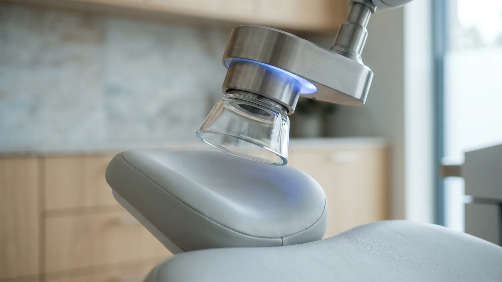 Nærbilde av dokkekjeglen på en femtosekundlaser for øyelaser, med et svakt blått lys, over en tom pasientstol i en lys nordisk øyeklinikk.