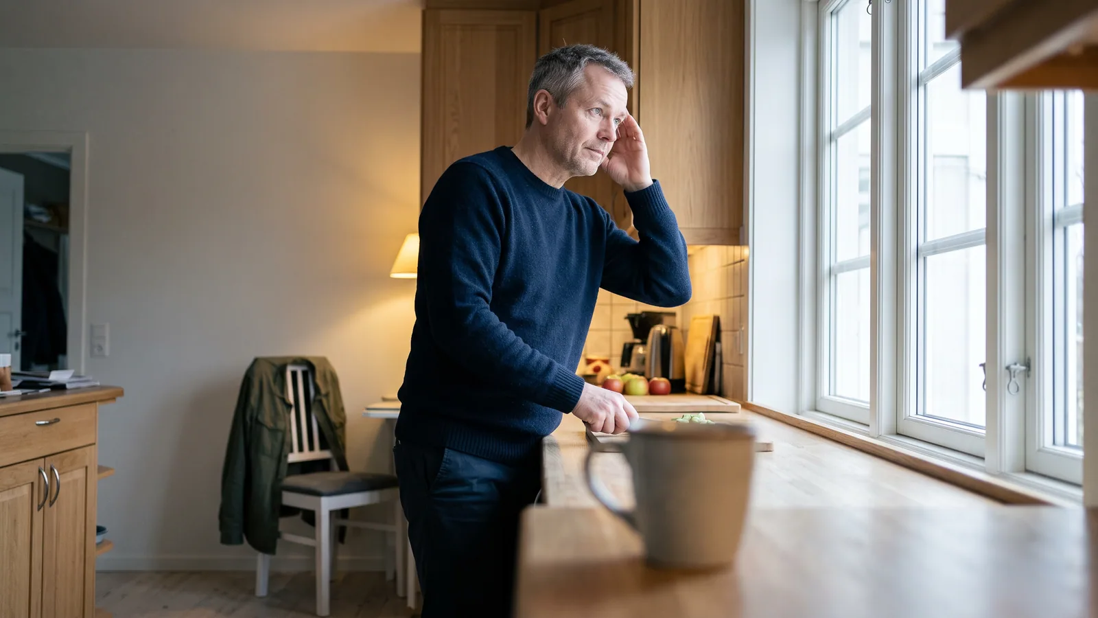 En mann i femtiårene står ved kjøkkenvinduet med hånden mot tinningen og et fjernt blikk ut i det grå dagslyset.